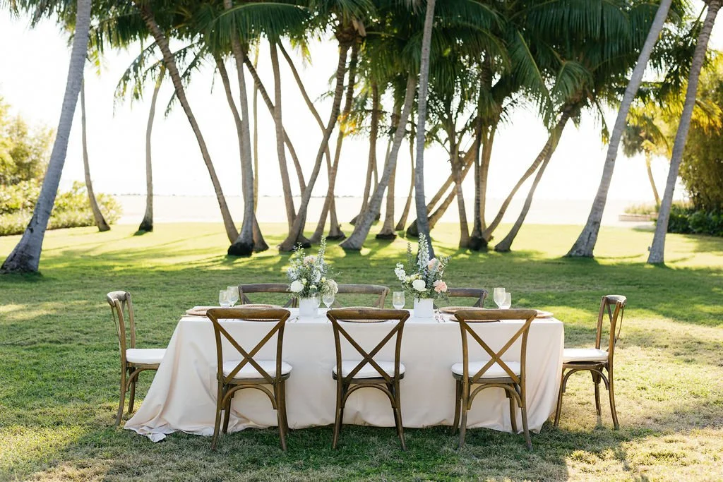 Outdoor dining table set with floral centerpieces and glasses, surrounded by wooden chairs, on lush green grass under tall palm trees overlooking a body of water at sunset.