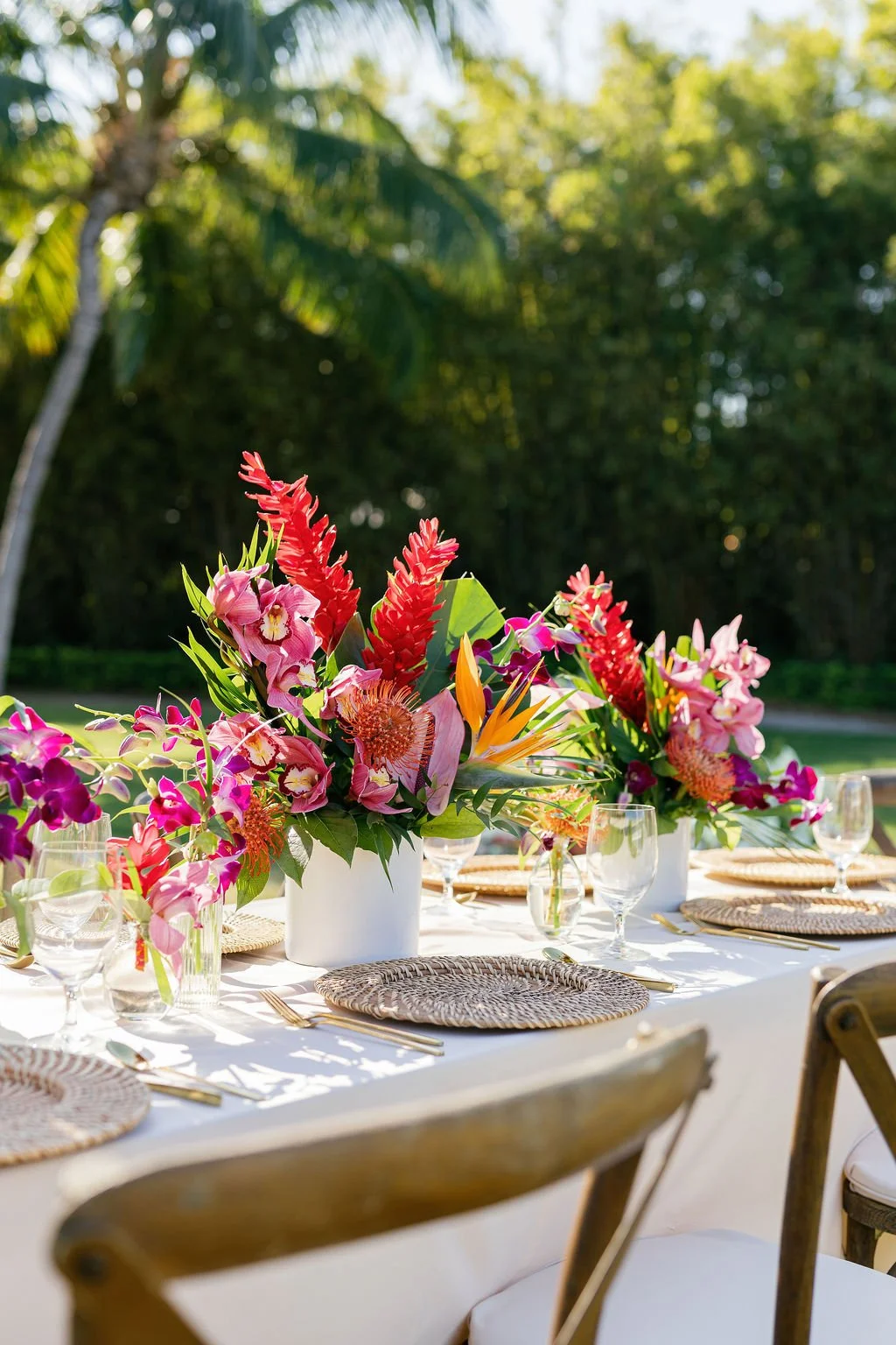 Colorful tropical flower arrangement on outdoor dining table with glassware and woven placemats, set in a lush garden during daytime.