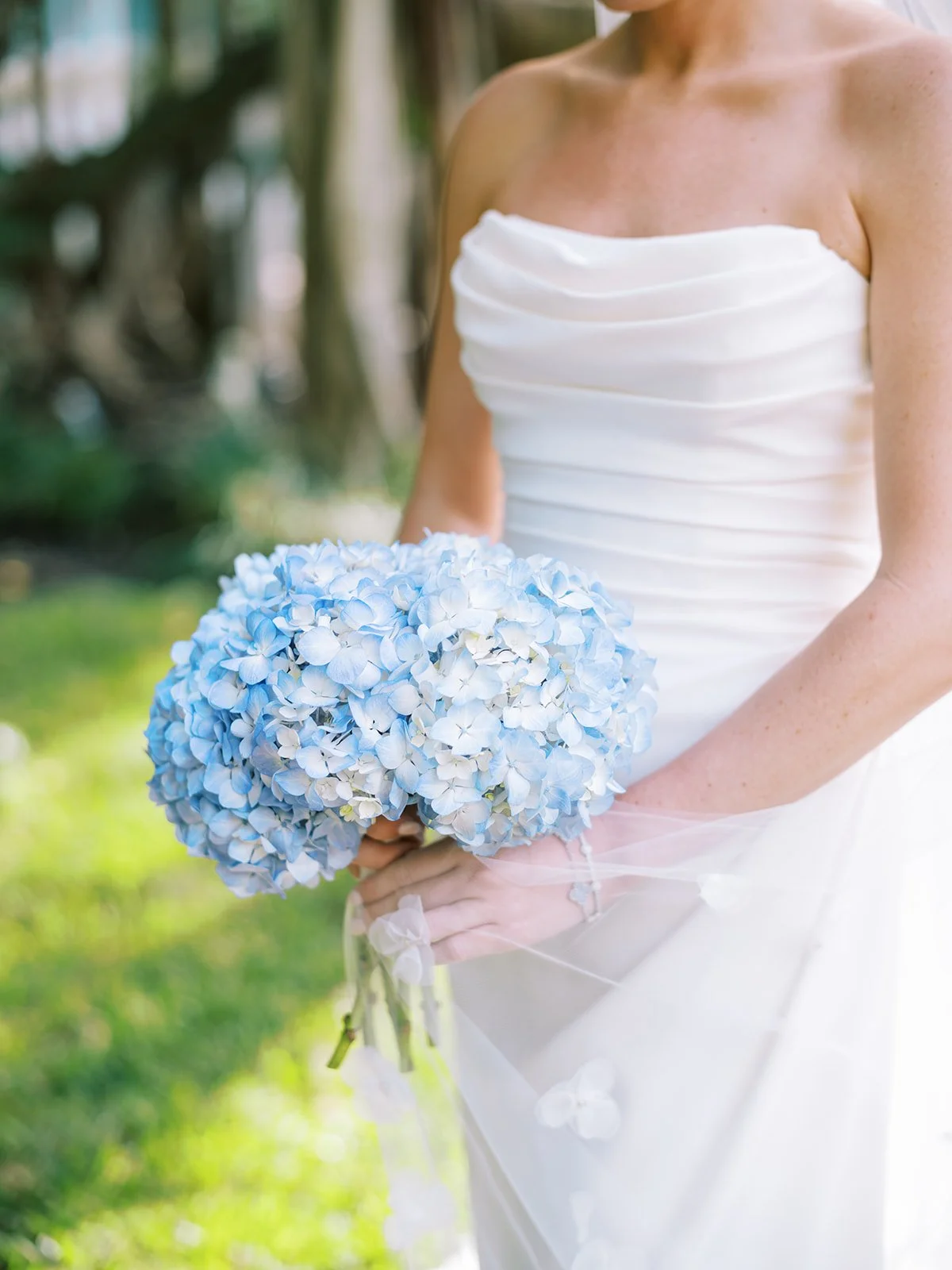 Bride holding a bouquet of blue hydrangeas while wearing a white strapless wedding dress.