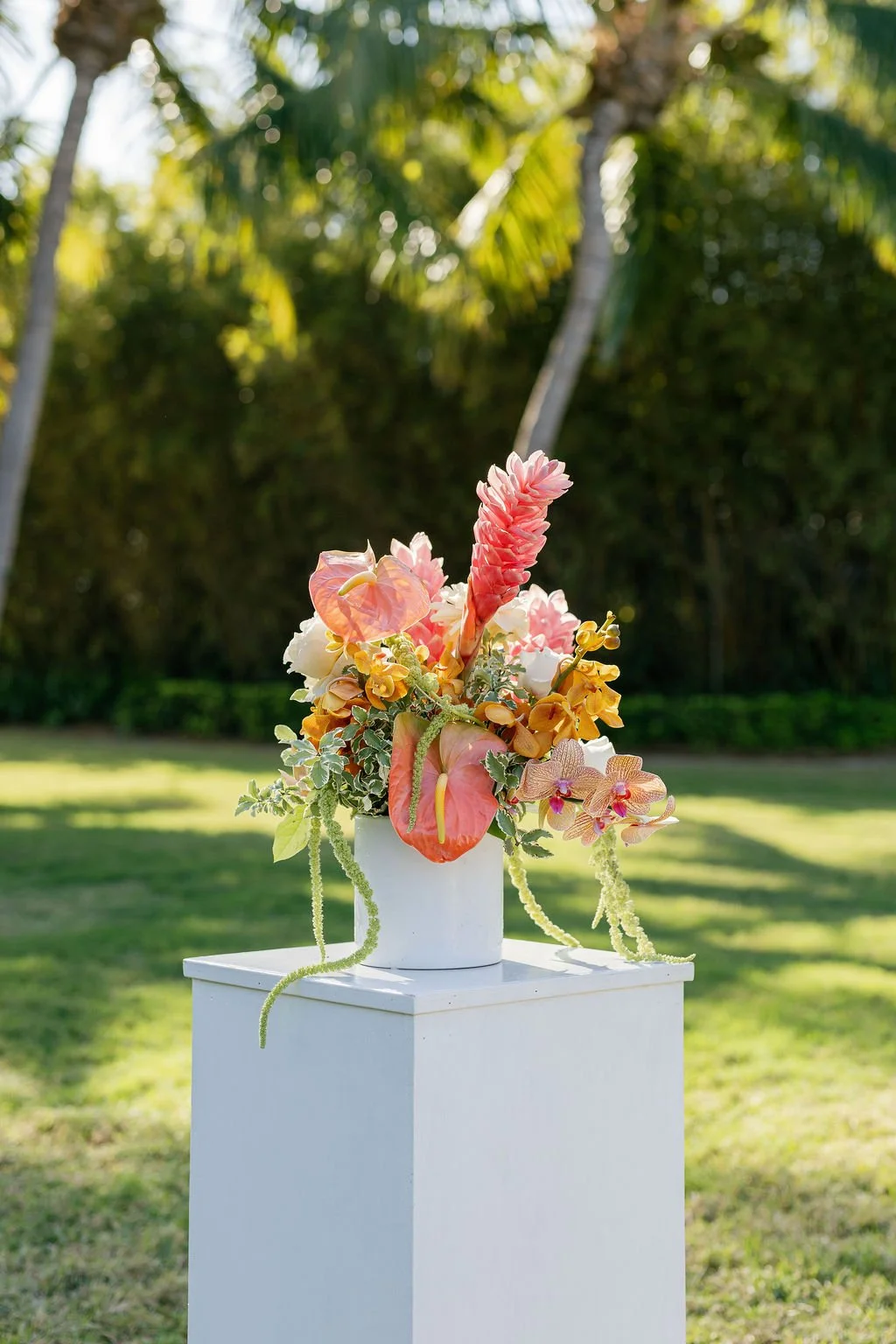 Colorful flower bouquet in a white vase on a white pedestal outdoors with greenery in the background.