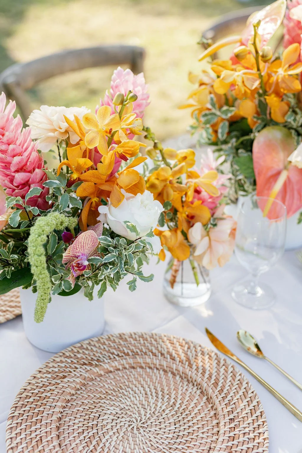 Colorful floral centerpiece with yellow, pink, white, and orange flowers on a white tablecloth, alongside gold cutlery and glassware.