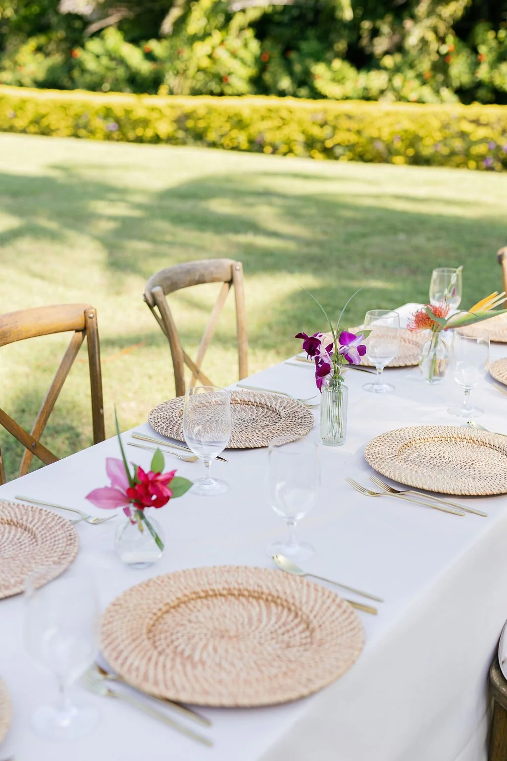 An outdoor dining table set with woven placemats, glassware, and colorful flowers in small vases, on a white tablecloth, with wooden chairs surrounding it, in a garden setting with green grass and trees.