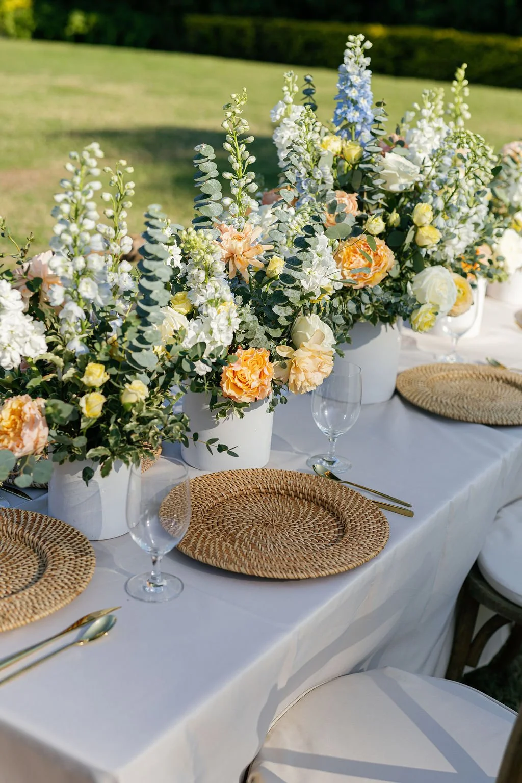 Elegant outdoor table setting with white floral arrangements, wicker placemats, clear wine glasses, and gold cutlery, set on a white tablecloth against a green lawn background.