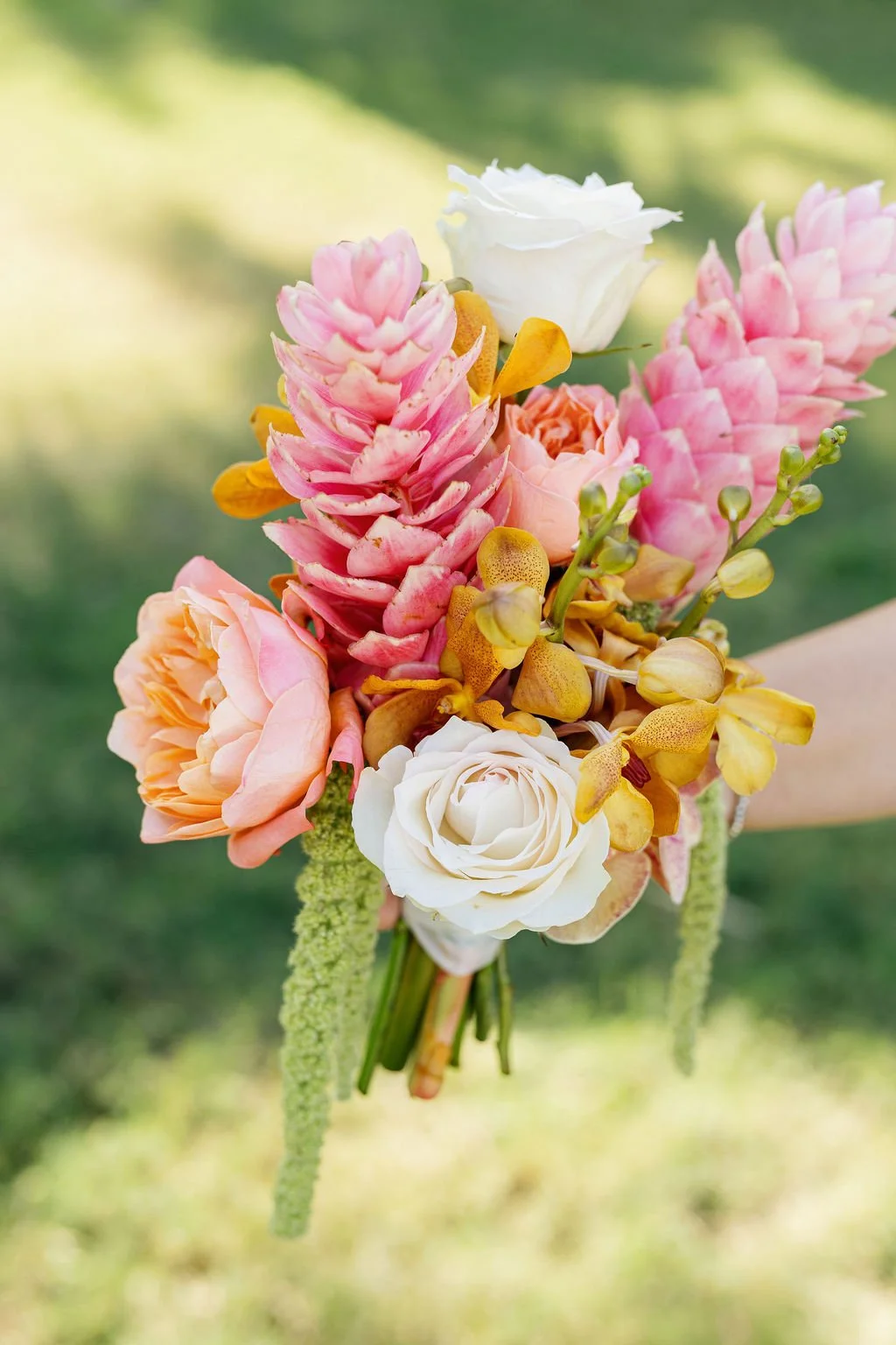 A bouquet of pink, white, peach, and yellow flowers held outdoors with a blurred green background.