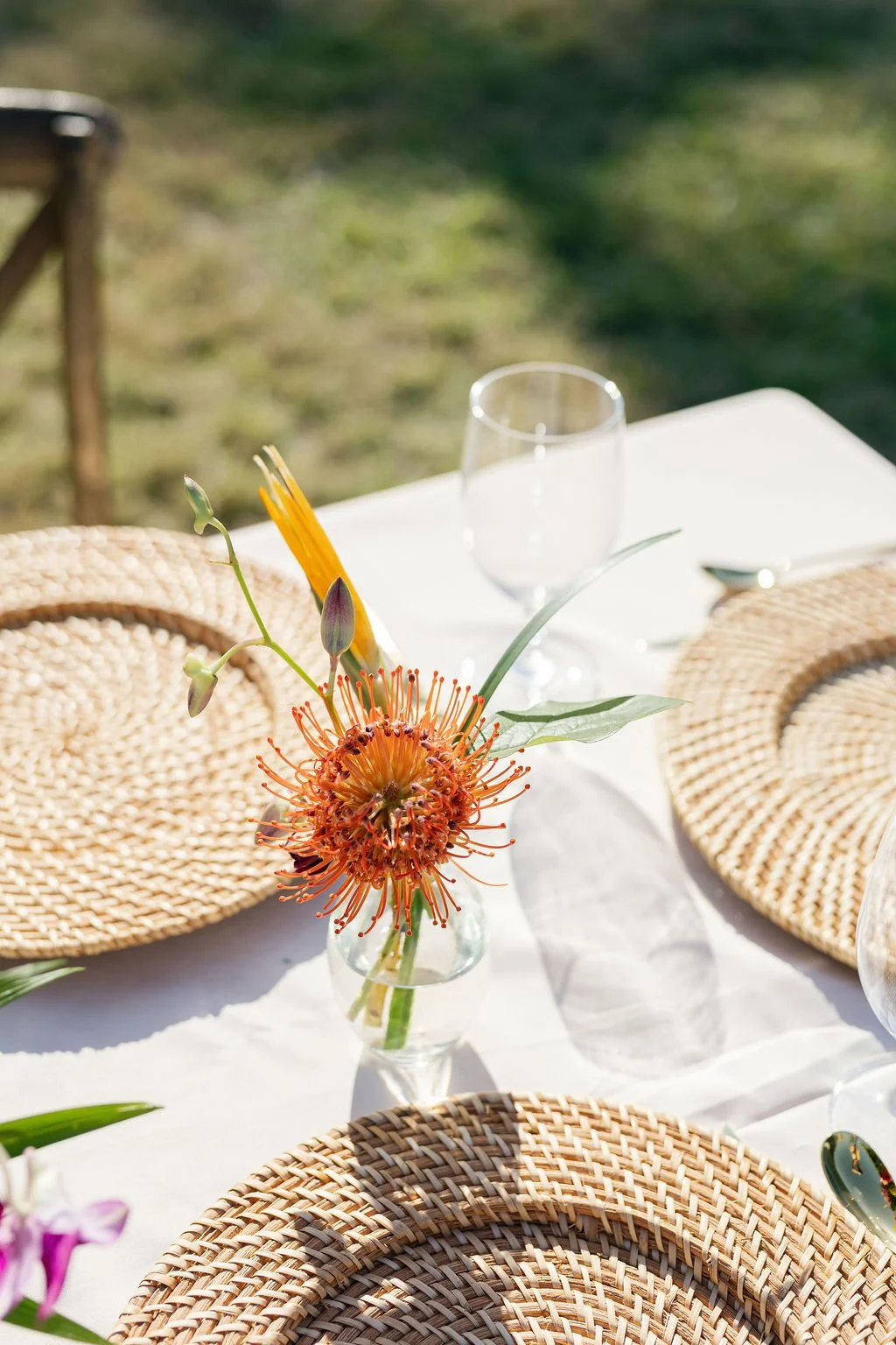 A table setup outdoors with a white tablecloth, woven placemats, a glass vase with orange flowers, and an empty wine glass.