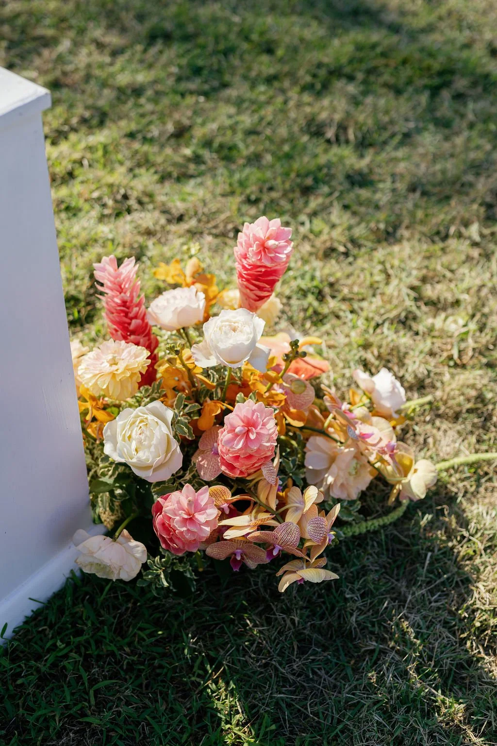 A colorful flower arrangement with pink, white, yellow, and peach flowers resting on a grassy ground next to a white monument.