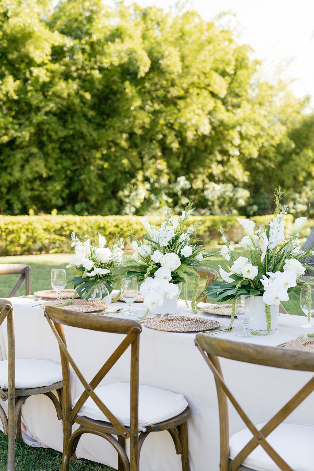 Outdoor dining table with white floral centerpieces, woven placemats, glassware, and wooden chairs, set in a garden with green trees in the background.