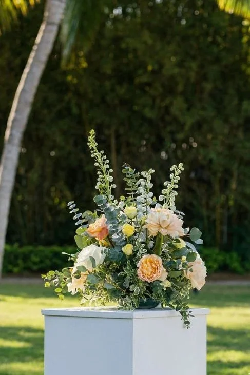 A floral arrangement in a white vase with white, blush, and peach-colored flowers, and green foliage, placed on a white pedestal outdoors.