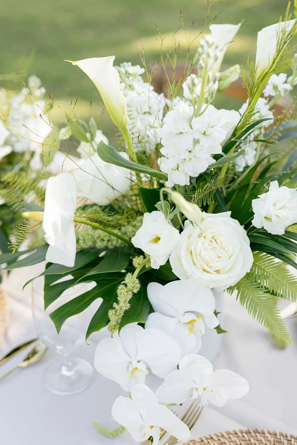 Close-up of a white floral centerpiece with roses, orchids, calla lilies, and greenery on a table with a blurred outdoor background.