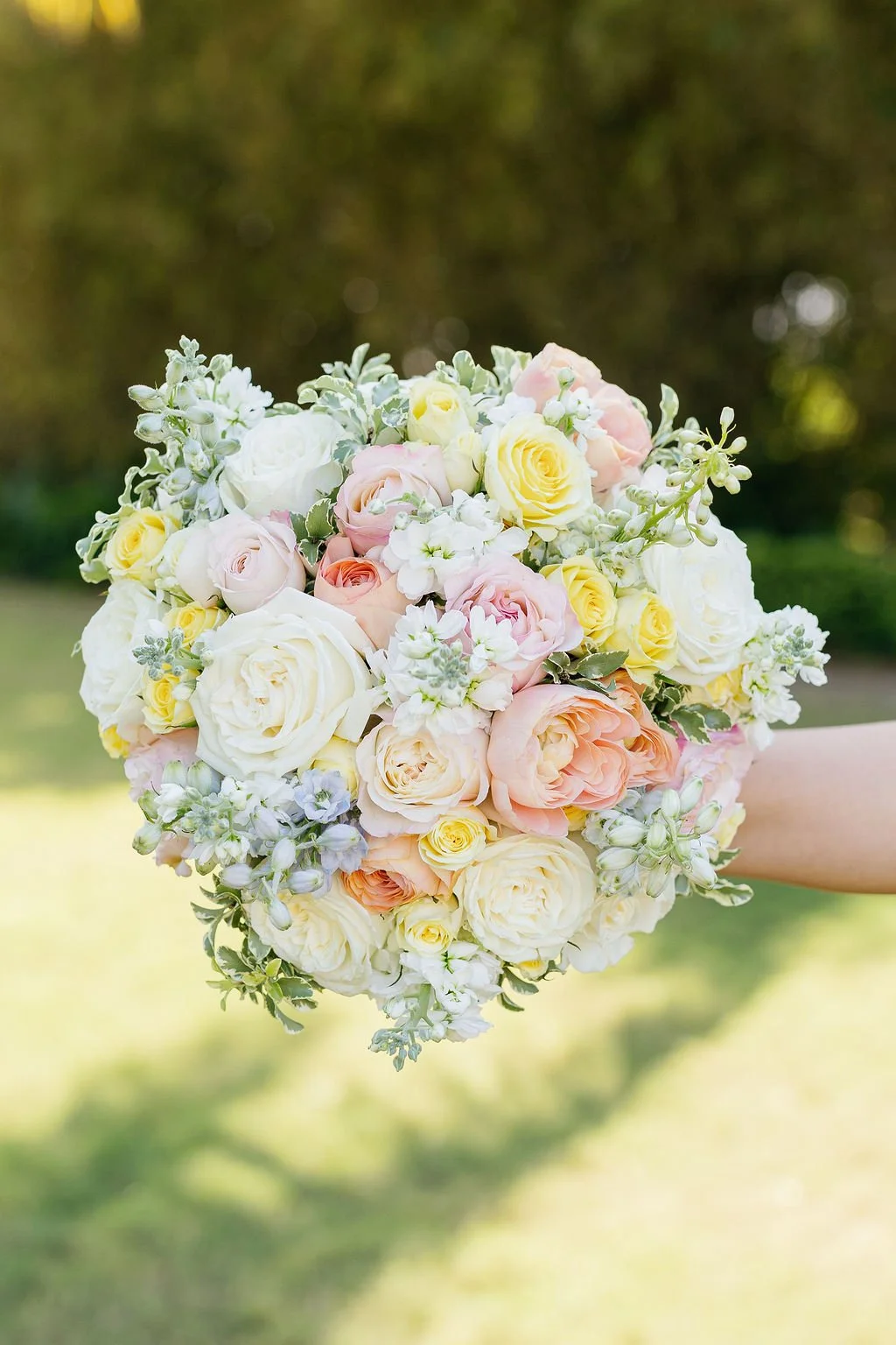 A round bouquet of pastel-colored roses and small white and purple flowers held outdoors on a sunny day.