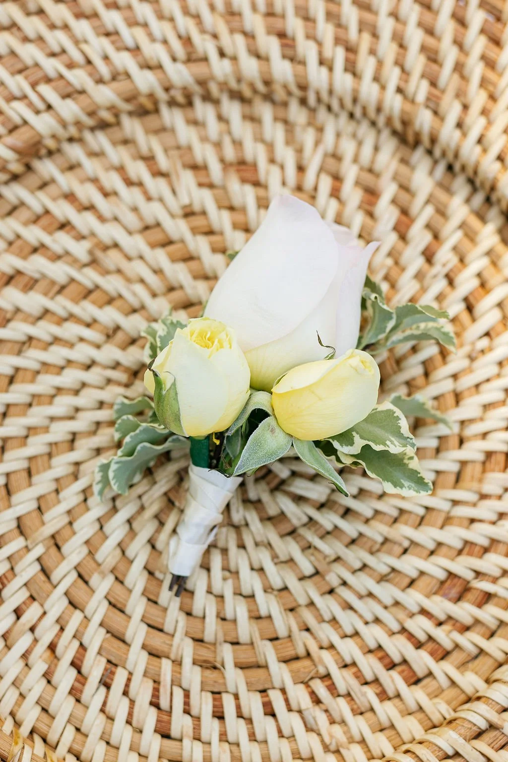 A white and yellow boutonniere with variegated green and white leaves on a woven rattan surface.