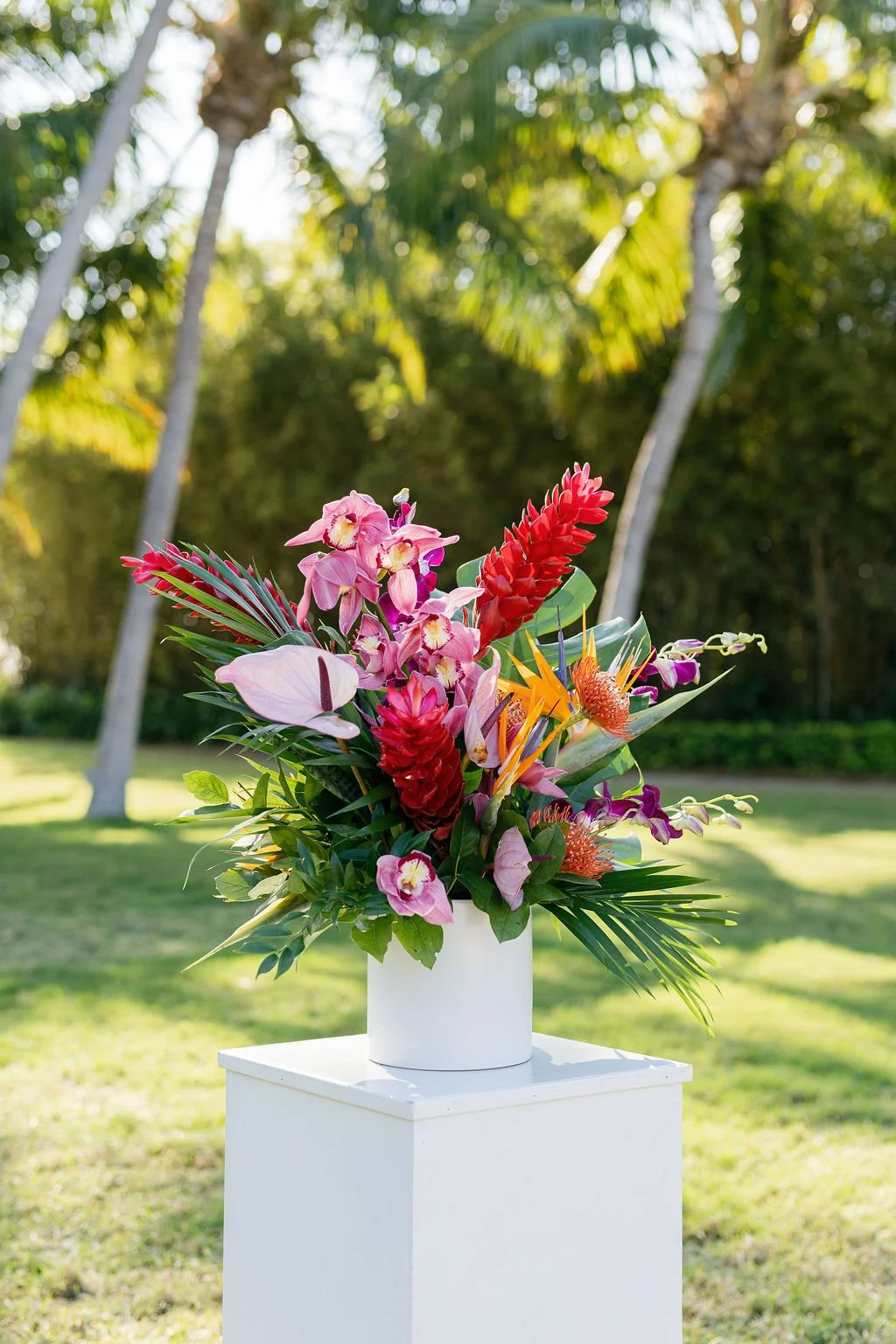 Colorful tropical flower arrangement in a white vase outdoors with palm trees and greenery in the background.