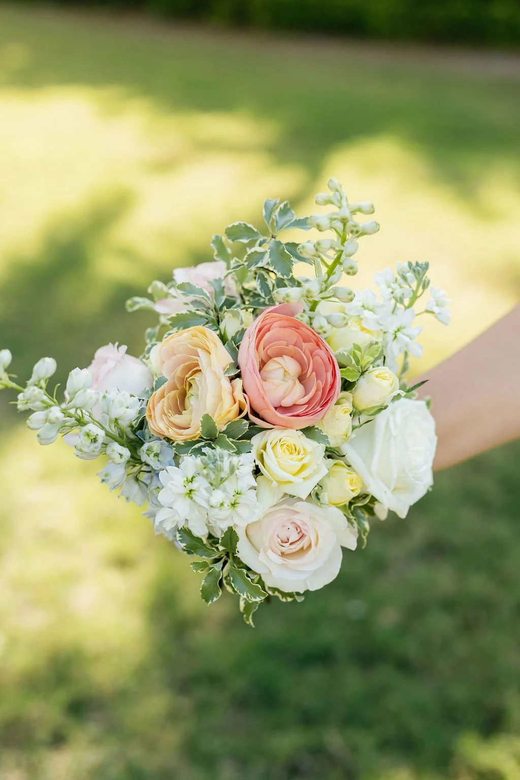 A hand holding a bouquet of flowers with white, yellow, and pink roses, and greenery against a blurred green background.