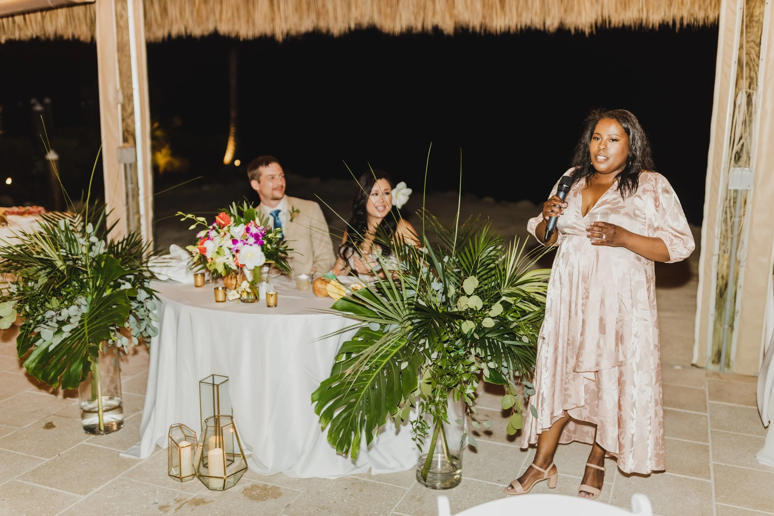 A woman gives a speech at a tropical-themed wedding reception. A couple, likely the bride and groom, sit at a decorated table with floral arrangements. Large green leaves in vases adorn the space, and candles are placed on the ground.