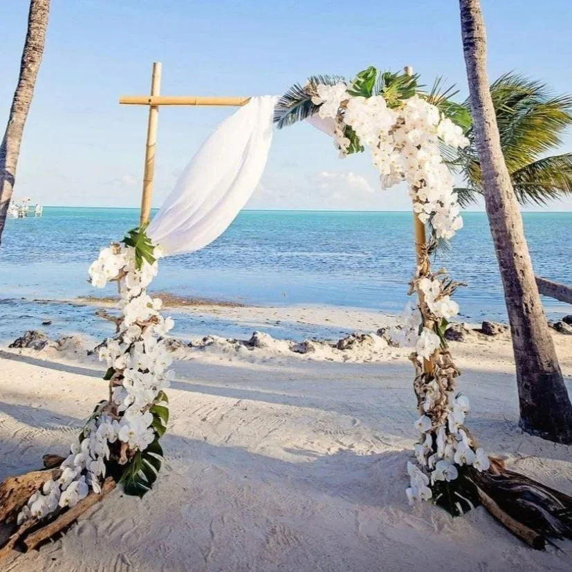 A wedding arch decorated with white flowers and fabric, set on a sandy beach with palm trees and ocean in the background.