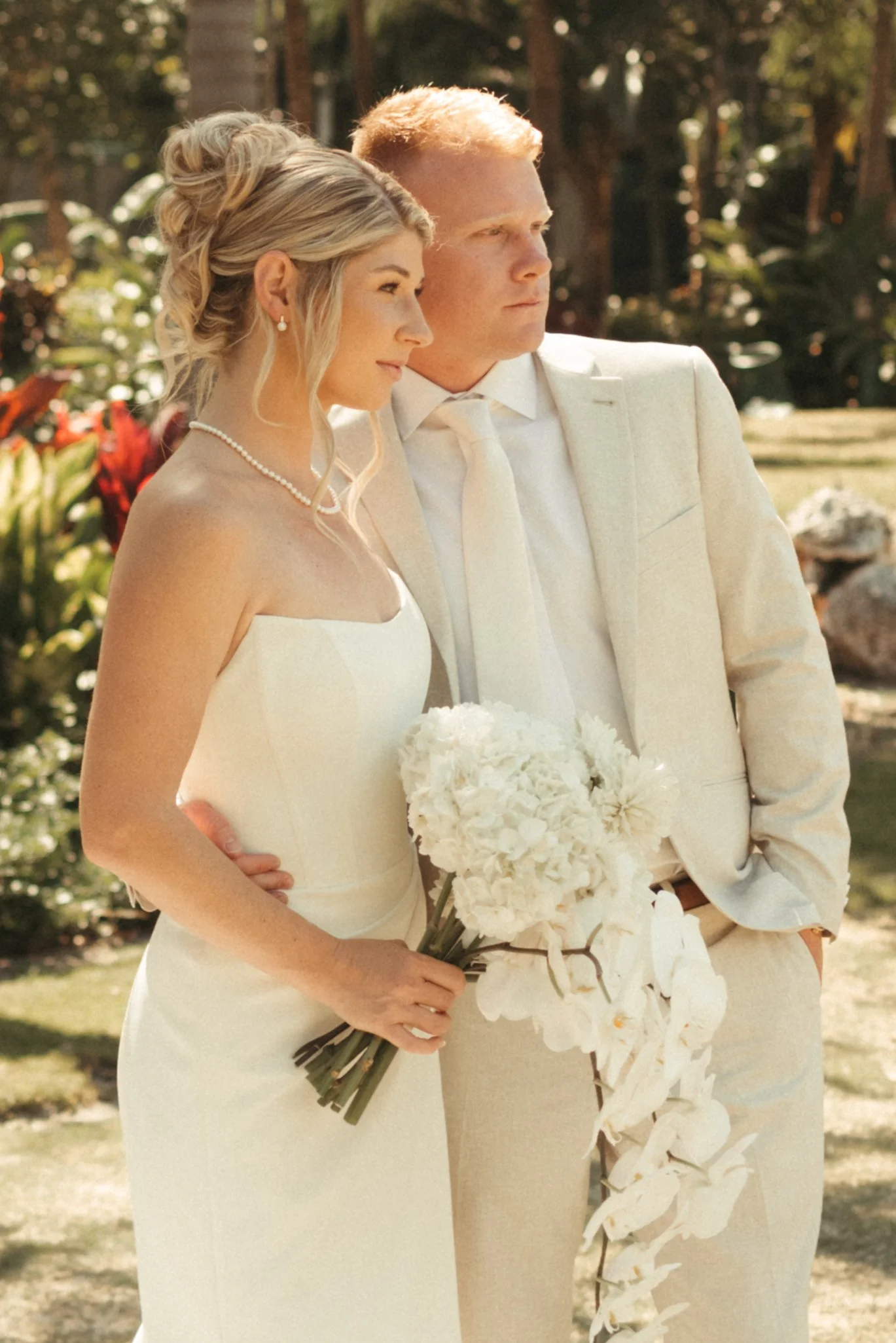 A bride and groom in formal attire, the bride holding a bouquet of white flowers, standing outdoors in a garden setting.