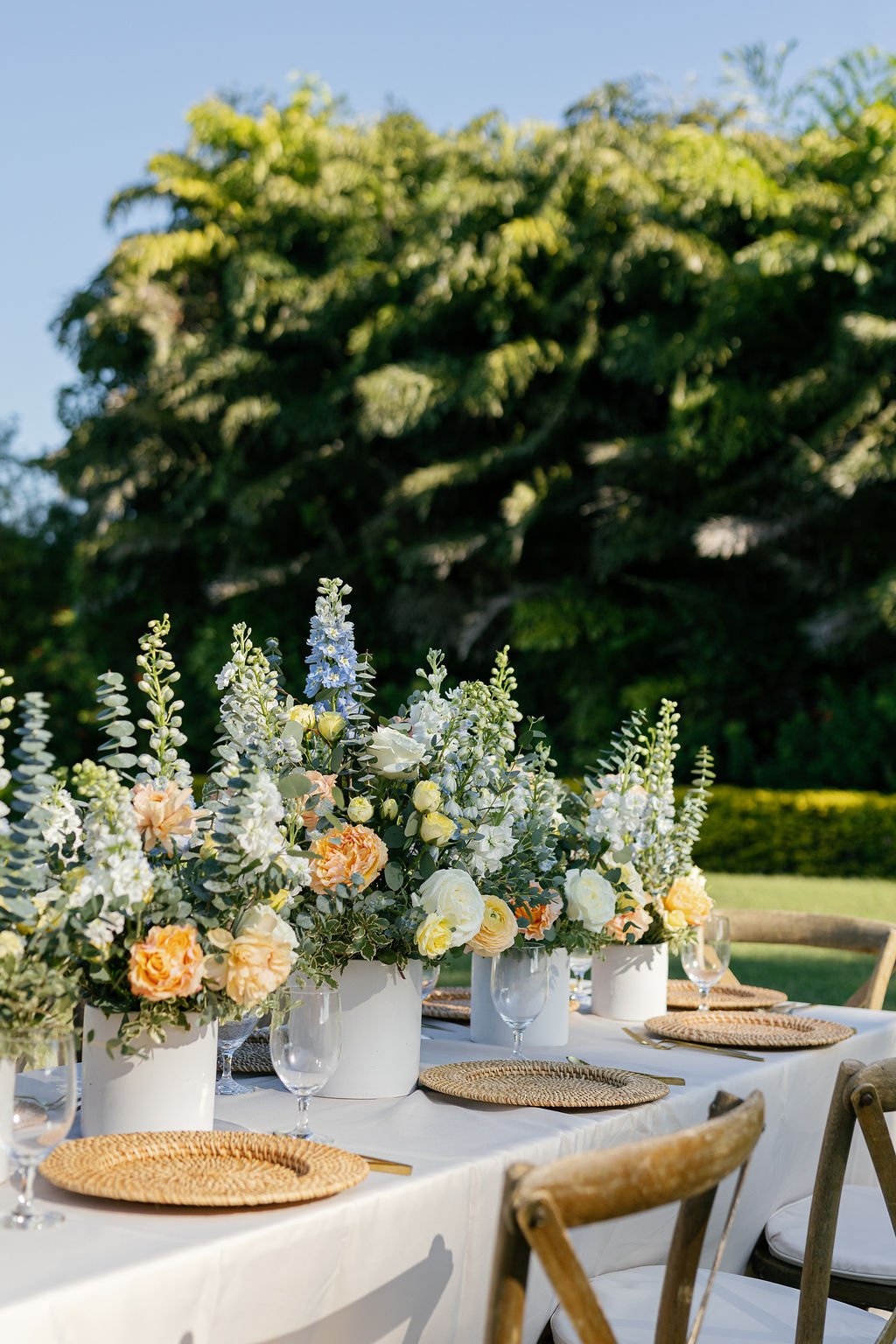 A table decorated with floral arrangements in white pots, set outdoors against a backdrop of green trees and a blue sky.