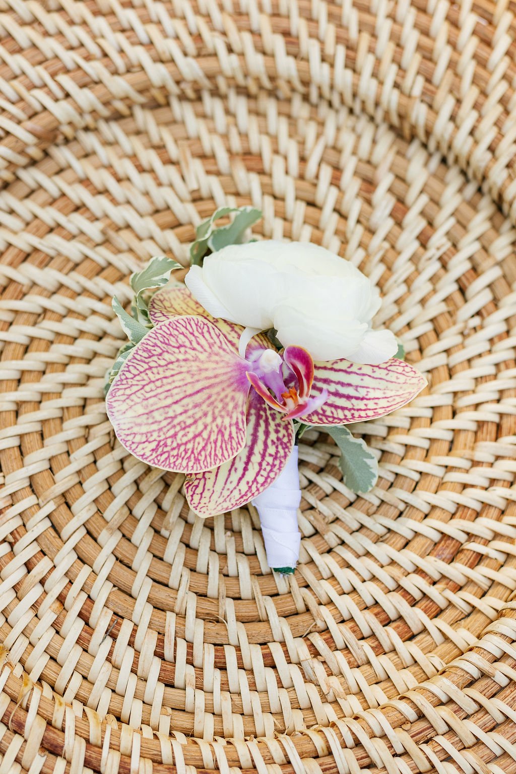 A boutonniere with a white flower, a pink and yellow patterned orchid, and green leaves, placed on a woven rattan surface.