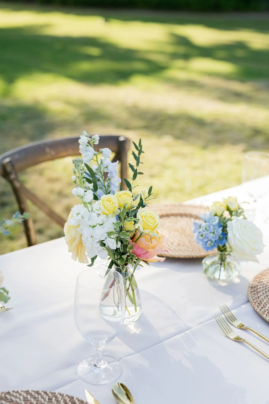 Floral arrangement with yellow roses, white flowers, and greenery in a glass vase on a white tablecloth.