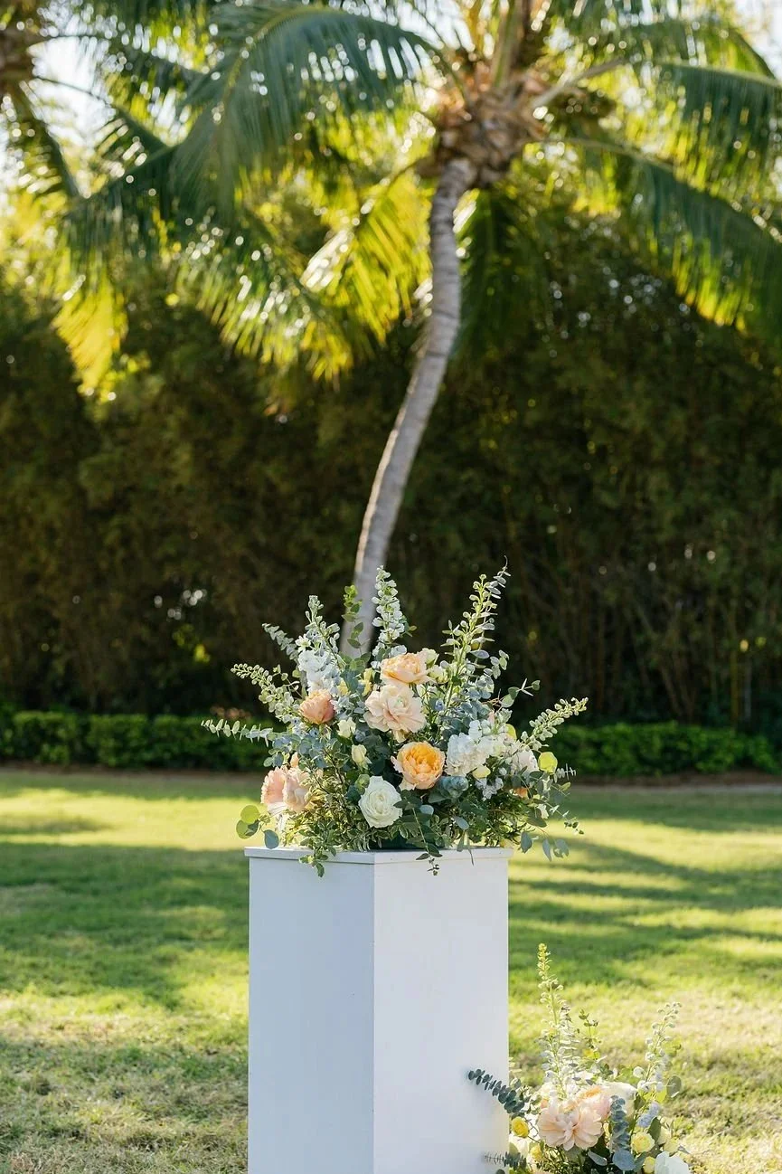 A floral arrangement featuring peach, white, and light purple flowers with greenery, placed on a white pedestal outdoors with a palm tree and green bushes in the background.