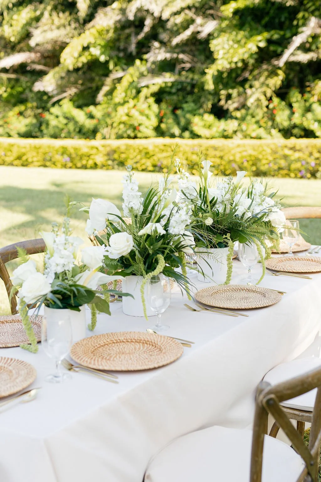 An outdoor dining table set with white floral centerpieces, wicker placemats, and glassware on a bright day in a garden.