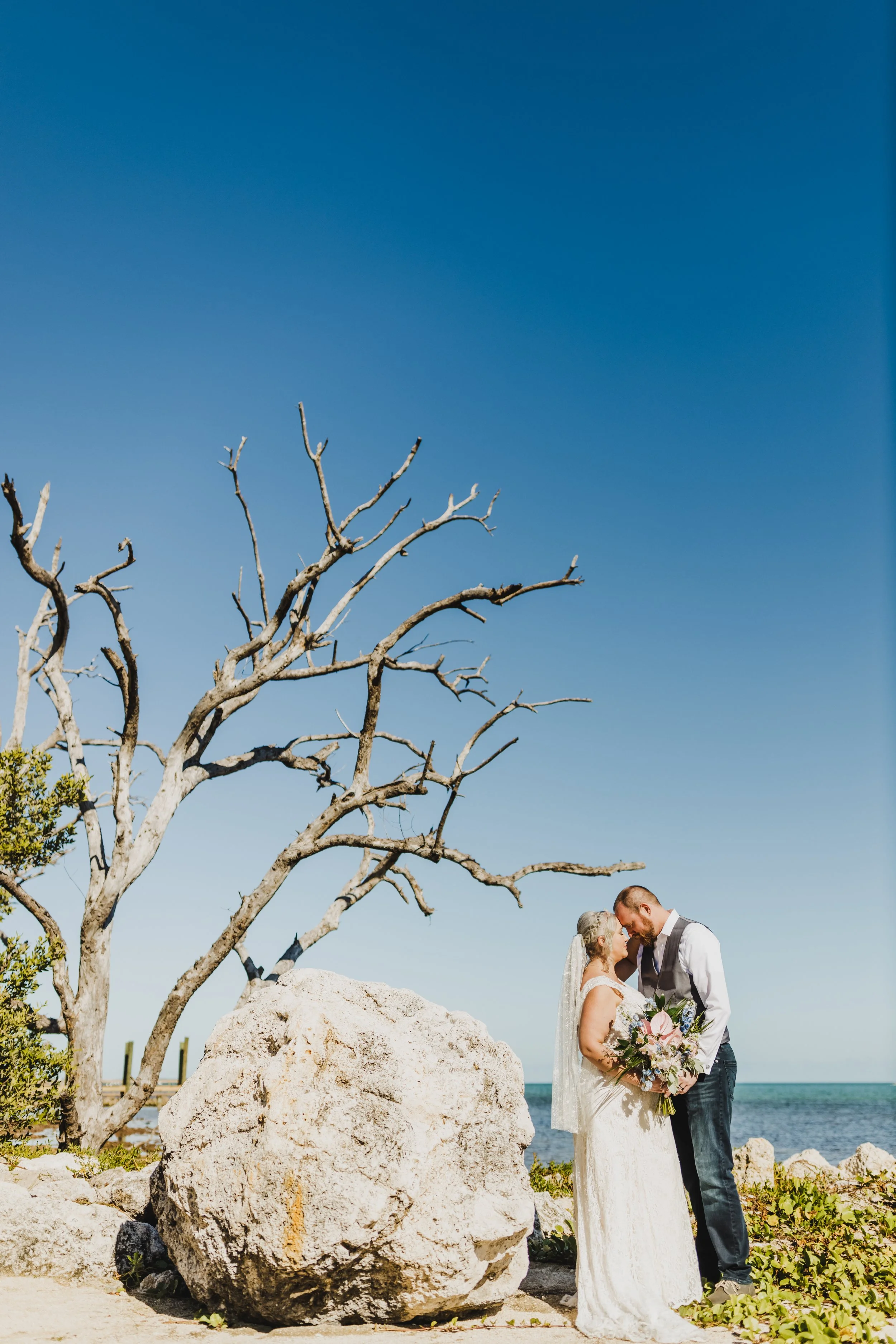 Bride and groom standing near a rocky shoreline with a bare tree and large boulder, under a clear blue sky.