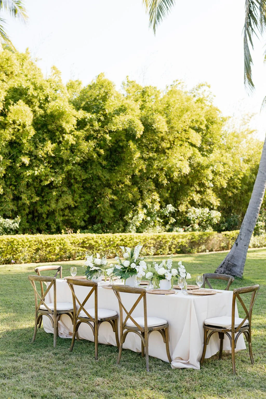 Outdoor wedding or event table setup with white tablecloth, floral centerpiece, and wooden chairs on grass, surrounded by greenery and trees.