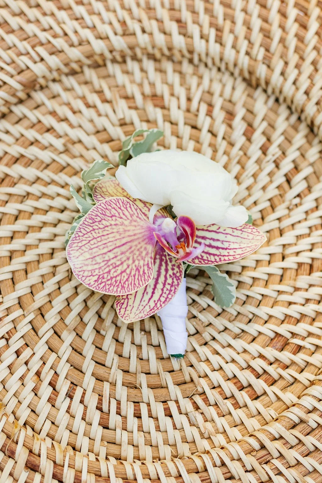 A boutonniere with a white flower, a pink and yellow striped orchid, green leaves, and purple-tinted foliage on a wicker basket background.