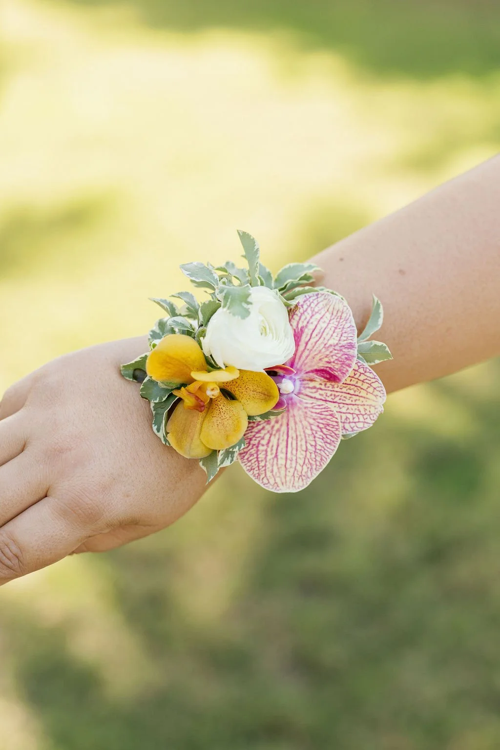 A hand wearing a colorful floral wrist corsage with yellow, white, and pink flowers against a blurred green background.