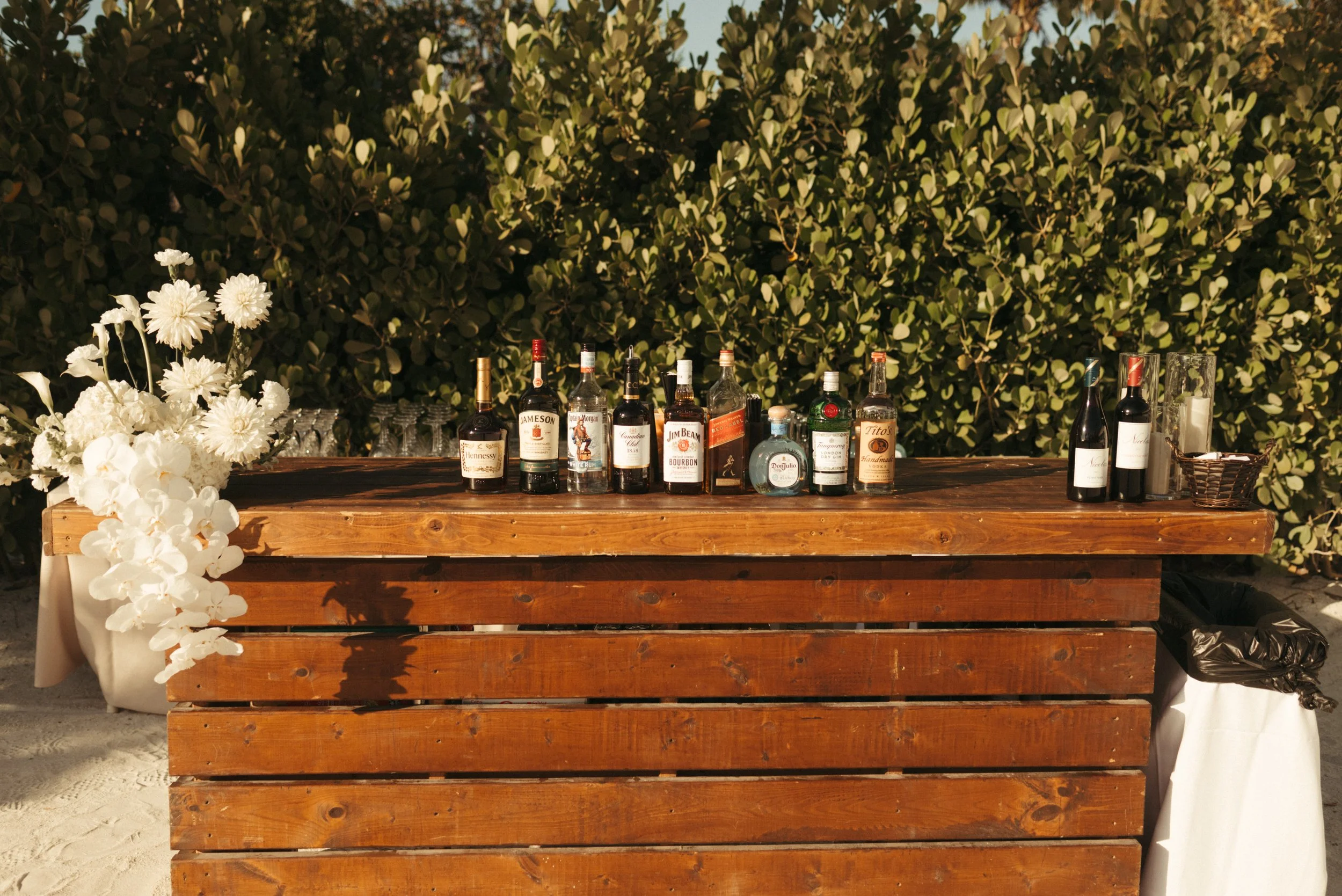 Outdoor bar setup with various liquor bottles and white flowers on a wooden counter.
