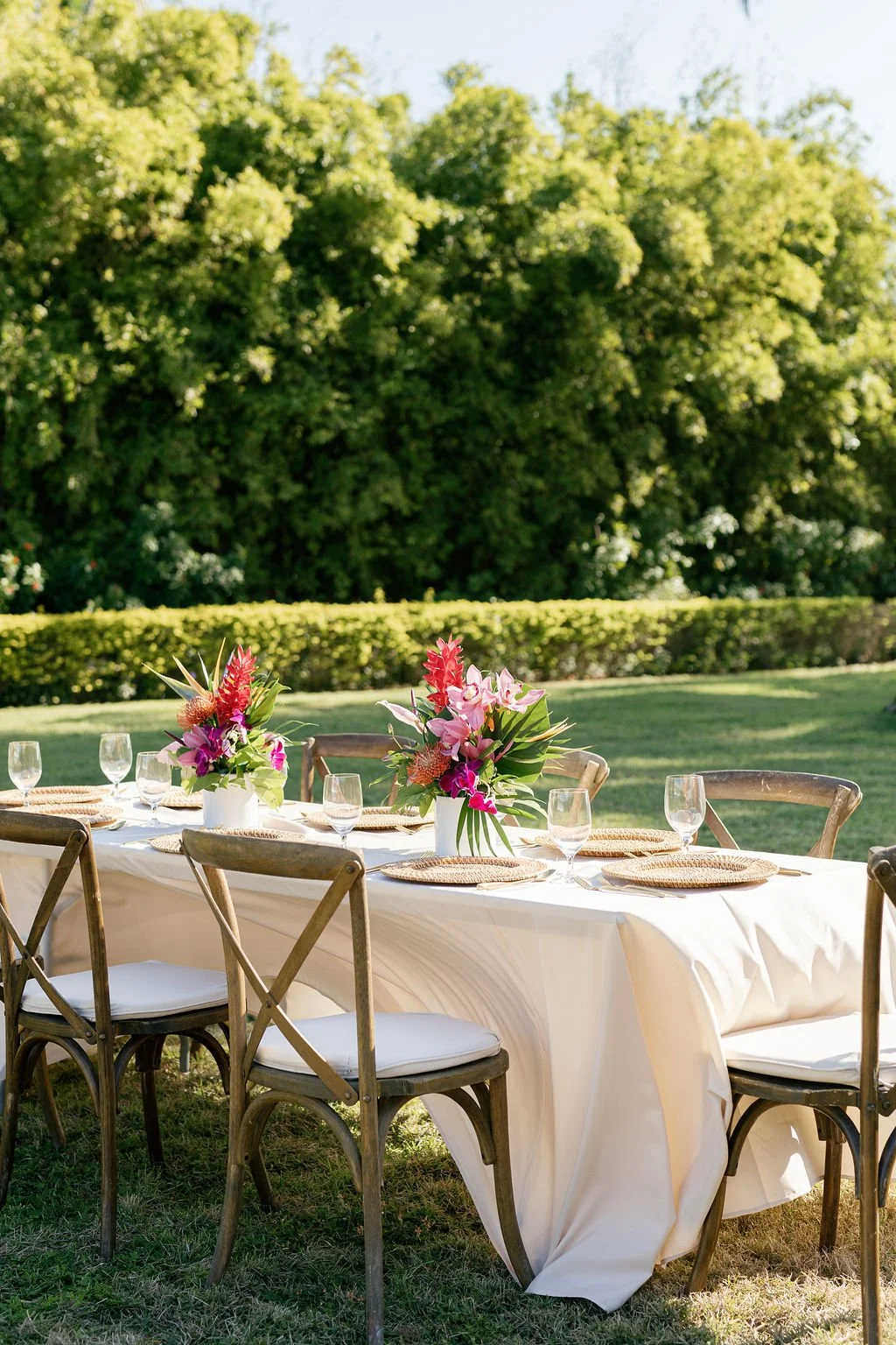 Outdoor table setup with white tablecloth, floral centerpieces, glassware, and woven placemats on a grassy area with trees in the background.