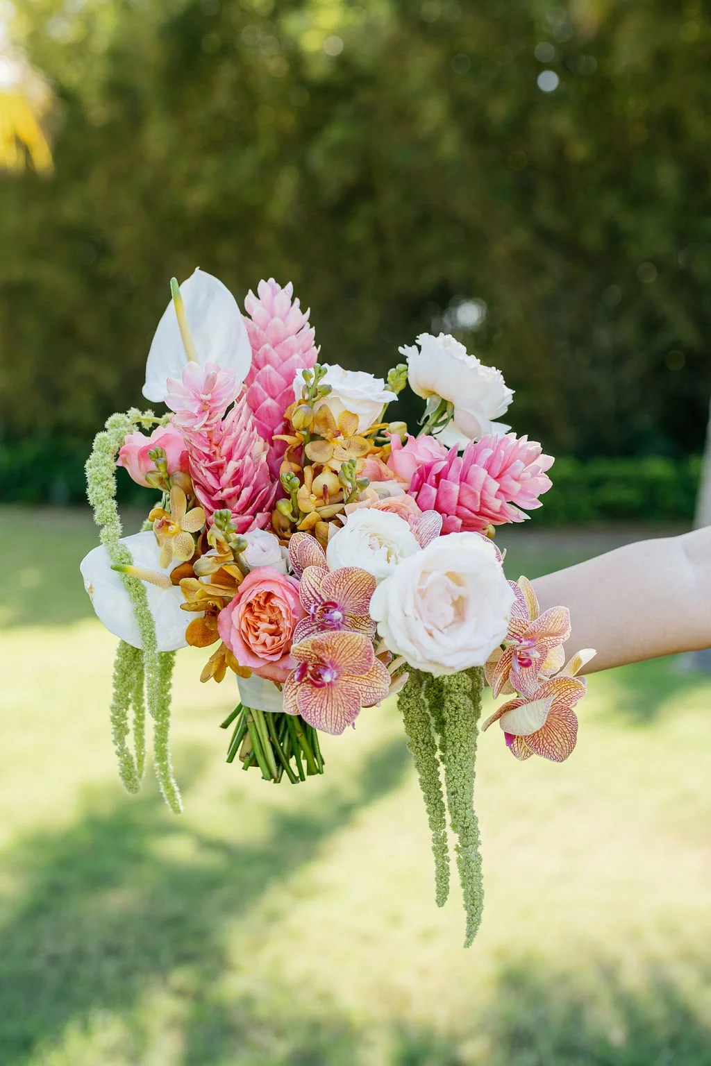 A colorful bouquet of various flowers held by a person outdoors. The bouquet includes white, pink, and peach roses, pink ginger flowers, orchids, and other greenery, with a blurred natural background.