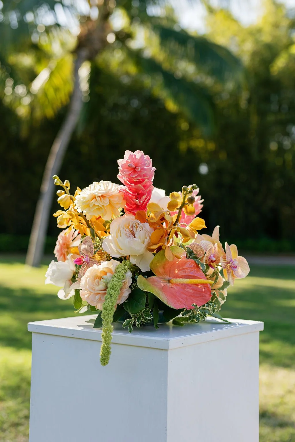 Colorful flower arrangement with pink, white, yellow, and peach flowers on a white pedestal outdoors, with trees and sunlight in the background.