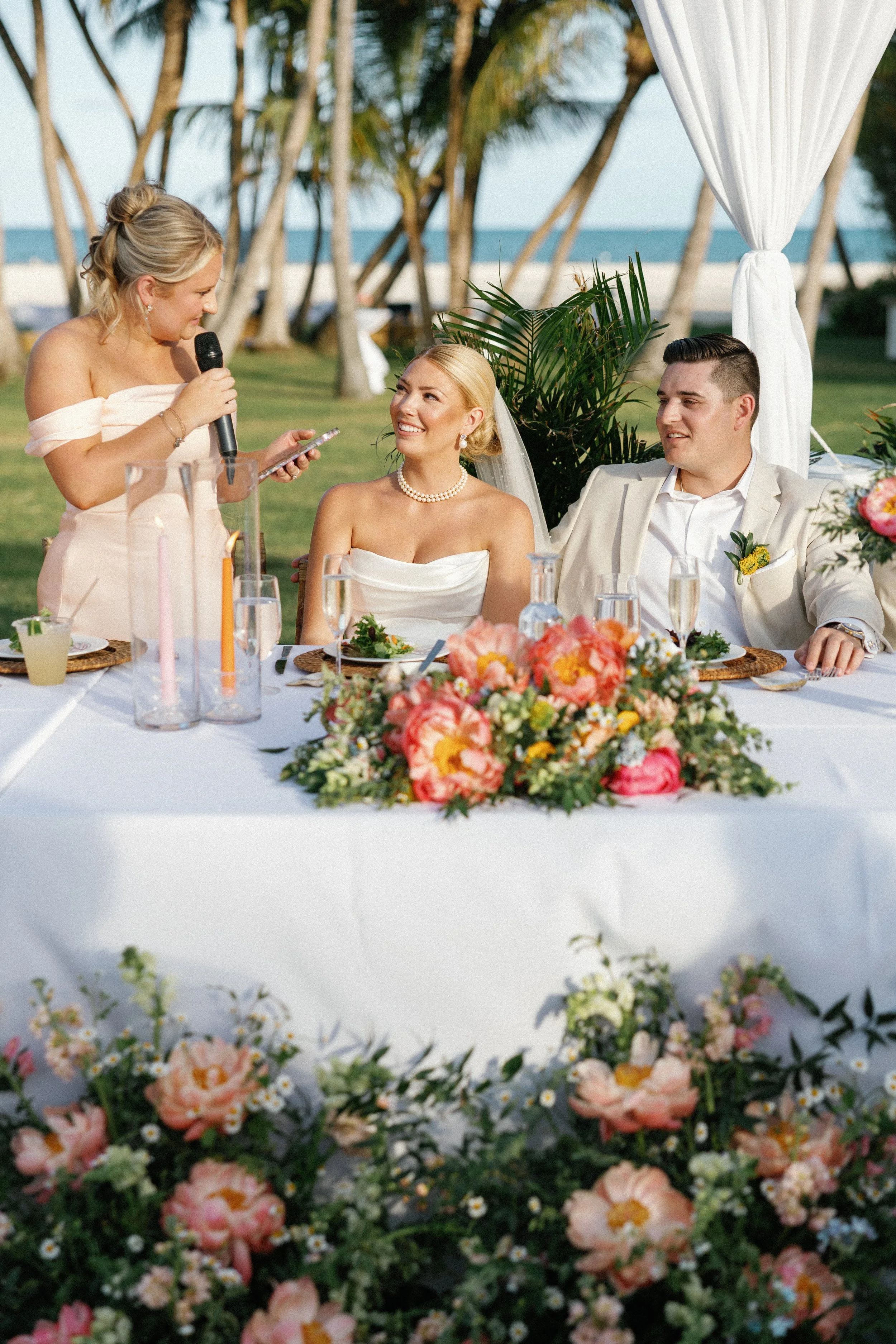 A wedding reception outdoors with a bride, groom, and a woman giving a speech. The table is decorated with a floral arrangement, and the background features palm trees, grass, and the ocean.