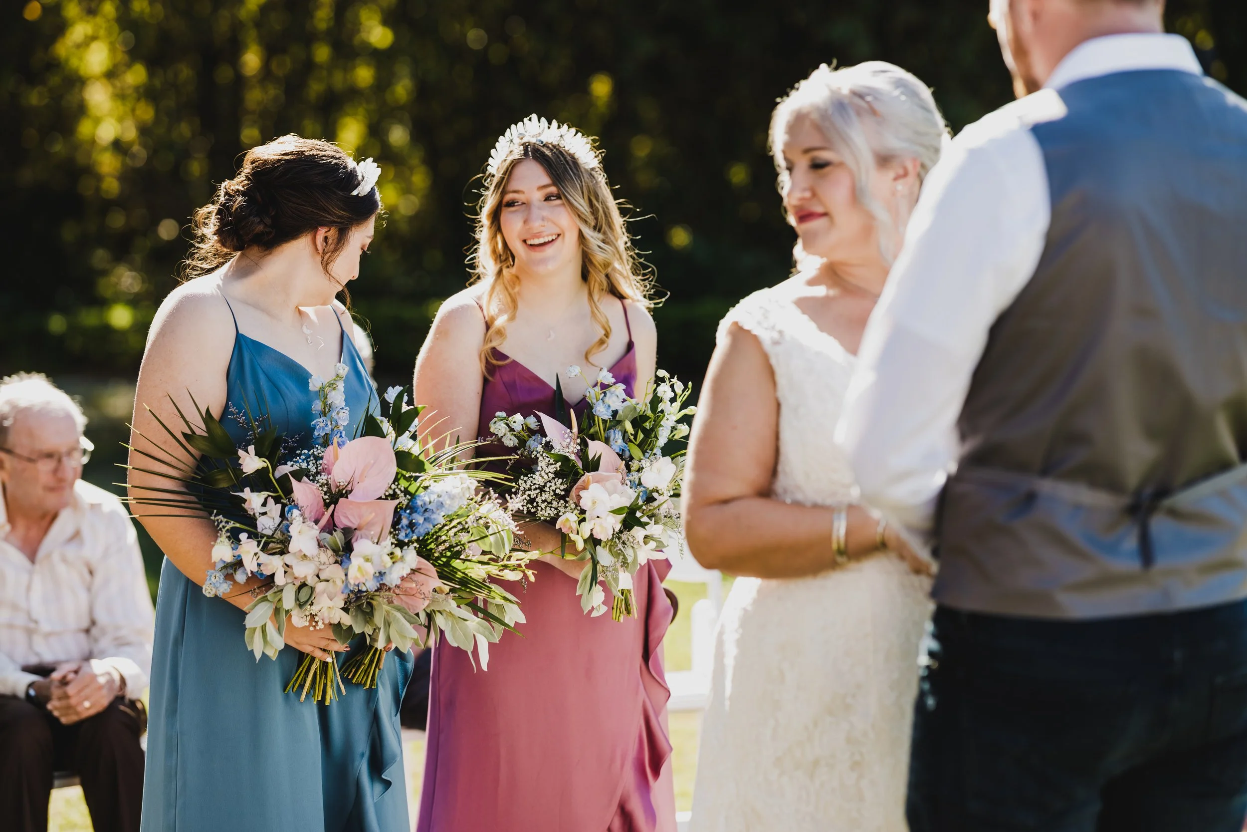 Bridal party with two women in blue and pink gowns holding bouquets, woman in white dress, and a man in a vest outdoors.