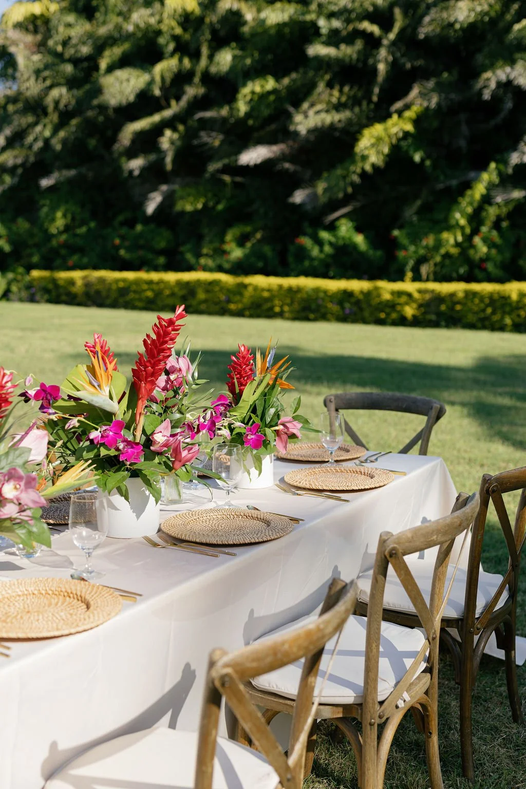 Outdoor dining table with floral centerpieces, wicker placemats, glasses, and cutlery, set on a lawn with trees and bushes in the background.