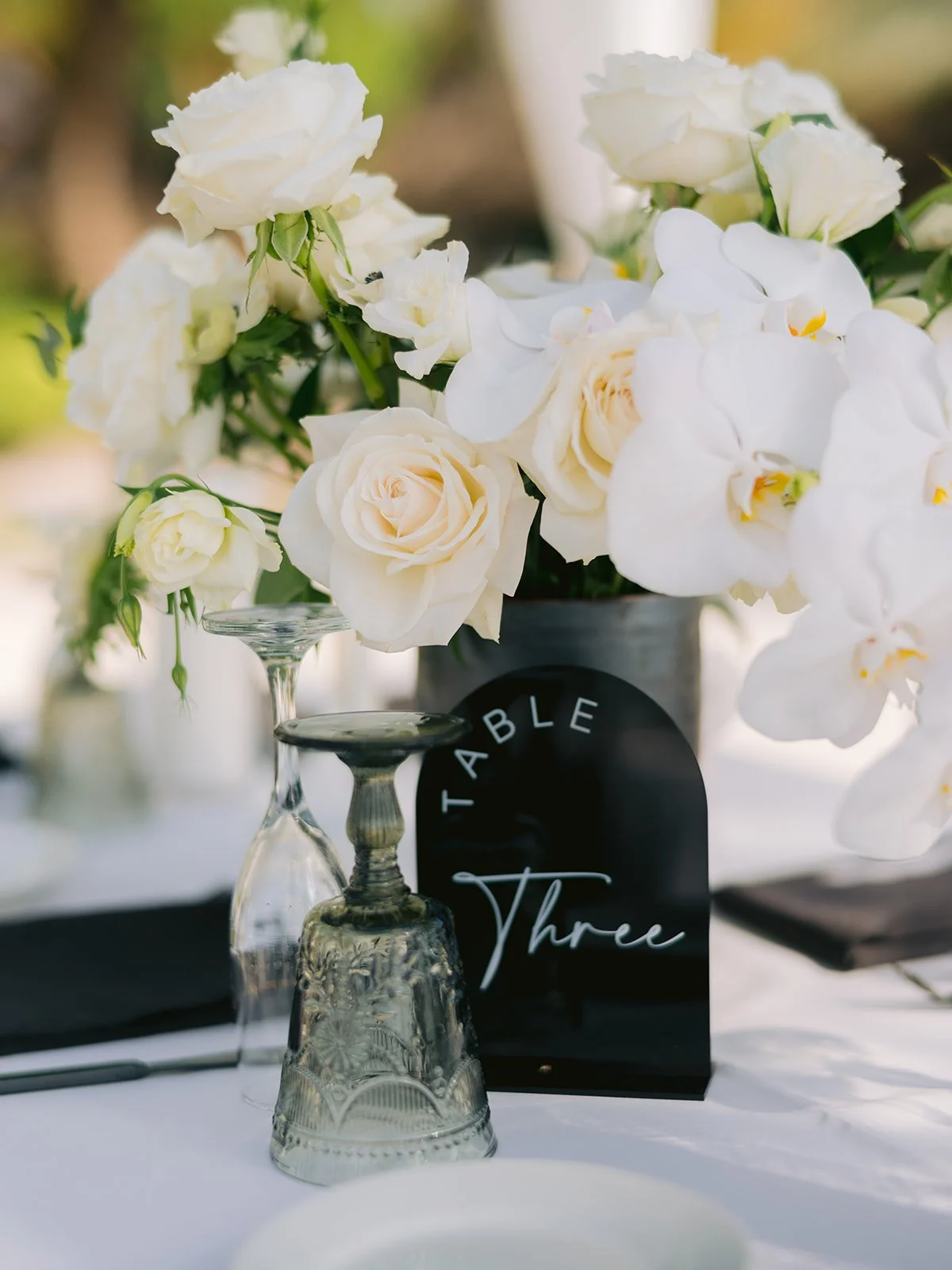 Table decorated with white flowers, including roses and orchids, a "Table Three" sign, and an inverted decorative glass.