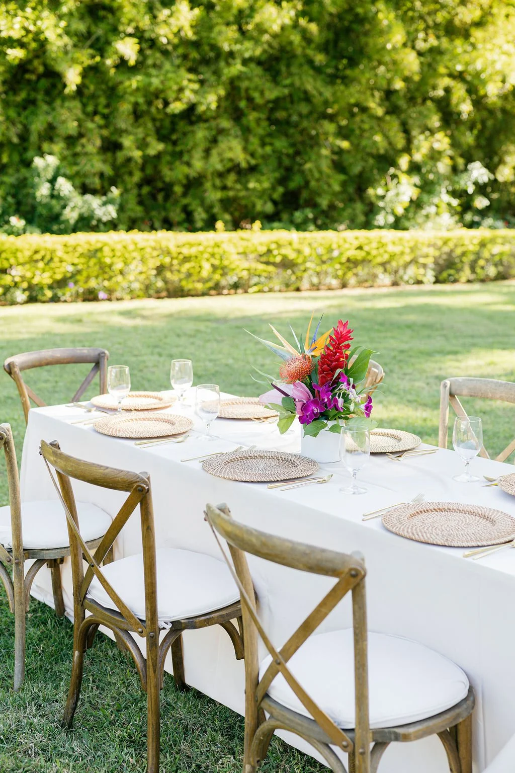 Outdoor dining table set with a white tablecloth, woven placemats, glassware, gold utensils, and a colorful floral centerpiece in a garden with green trees and grass.