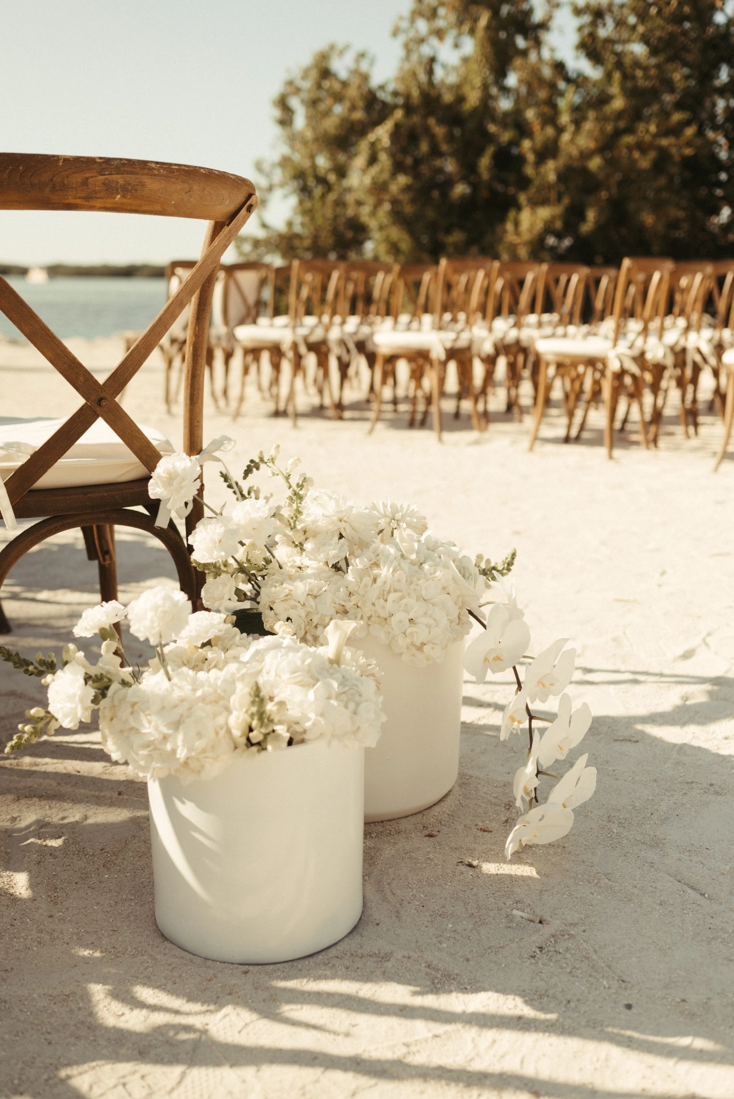 Outdoor wedding ceremony setup with wooden chairs and white flower arrangements in white vases on a sandy surface.