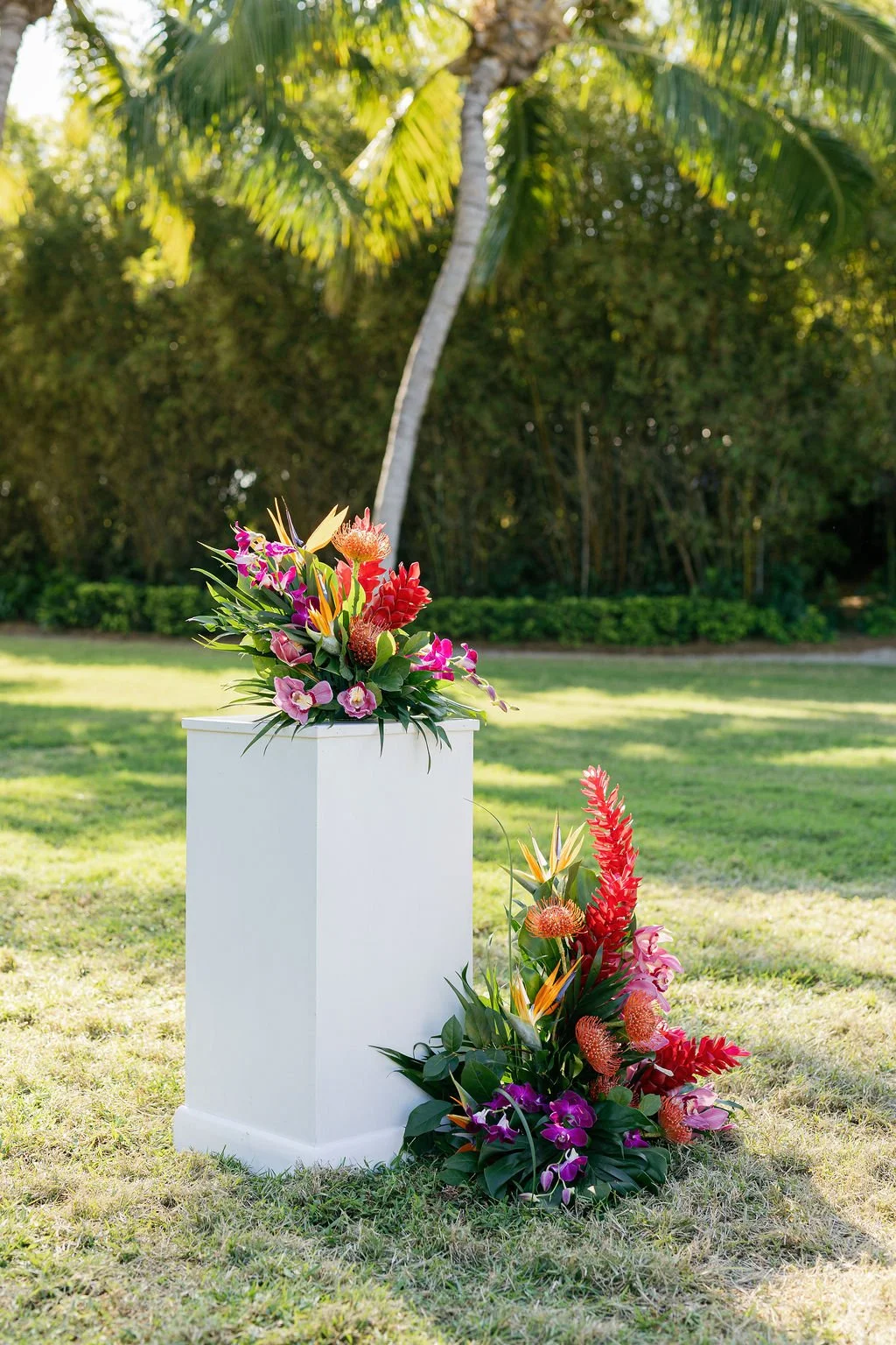 A white pedestal with a colorful floral arrangement featuring pink, purple, red, and yellow flowers, placed outdoors on grass with a background of trees and a palm tree.