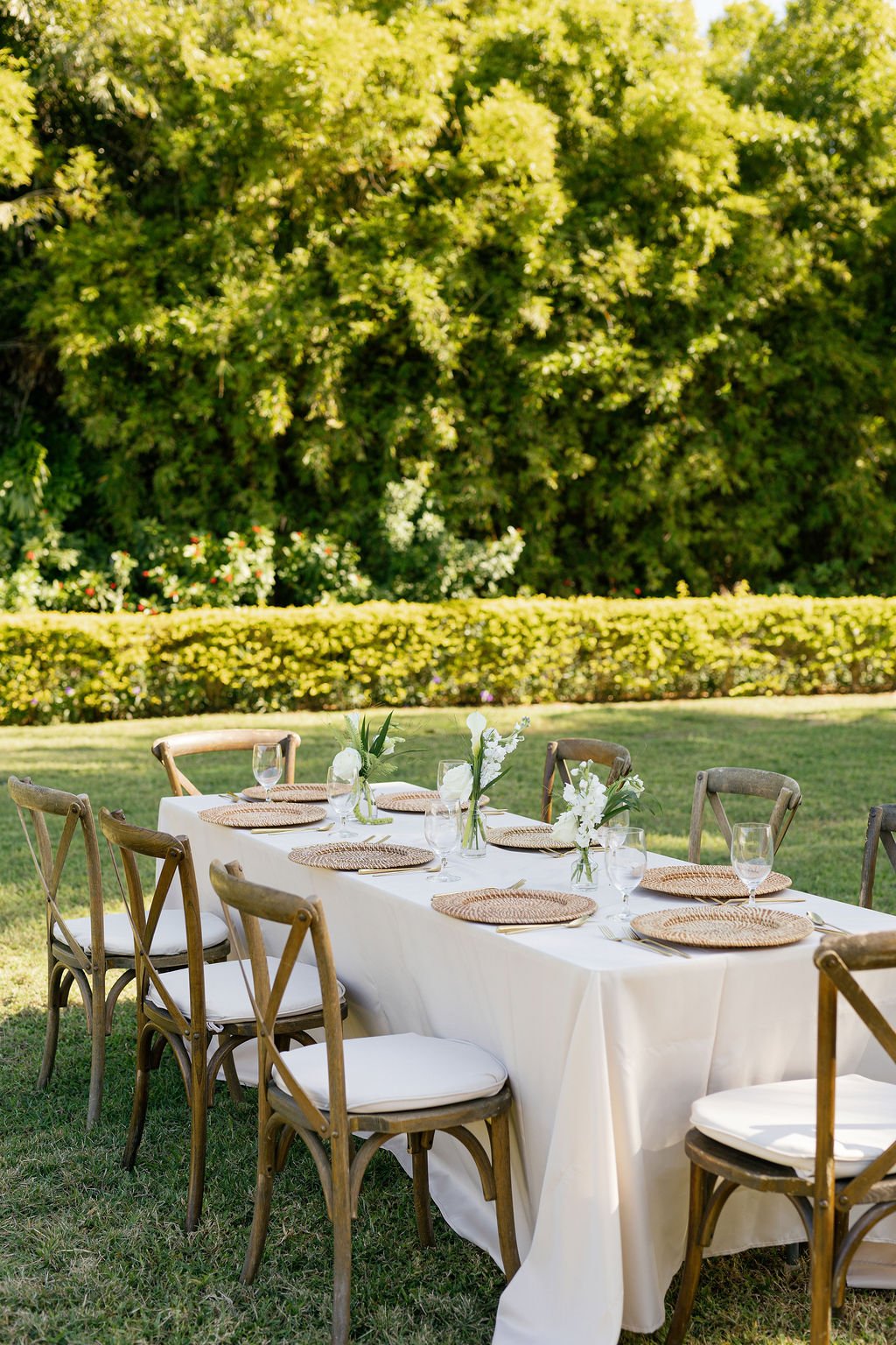 Outdoor dining table set for a gathering, with a white tablecloth, wicker placemats, glassware, and floral centerpieces, surrounded by wooden chairs, on a lawn with lush green trees and bushes in the background.