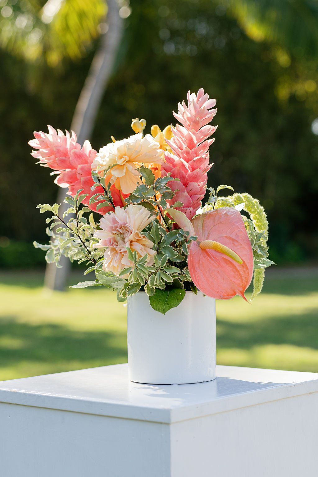 A floral arrangement with pink, cream, and peach flowers in a white vase on a white table outdoors, with greenery and trees in the background.