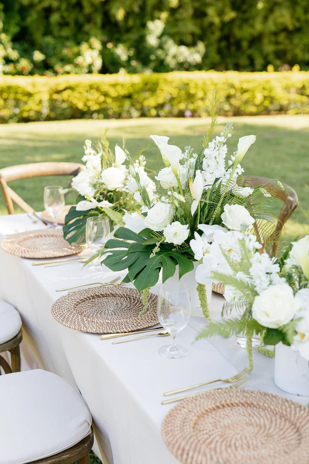 Elegant outdoor table setting with white flowers and greenery, gold flatware, woven placemats, and clear wine glasses, in a garden or park area.