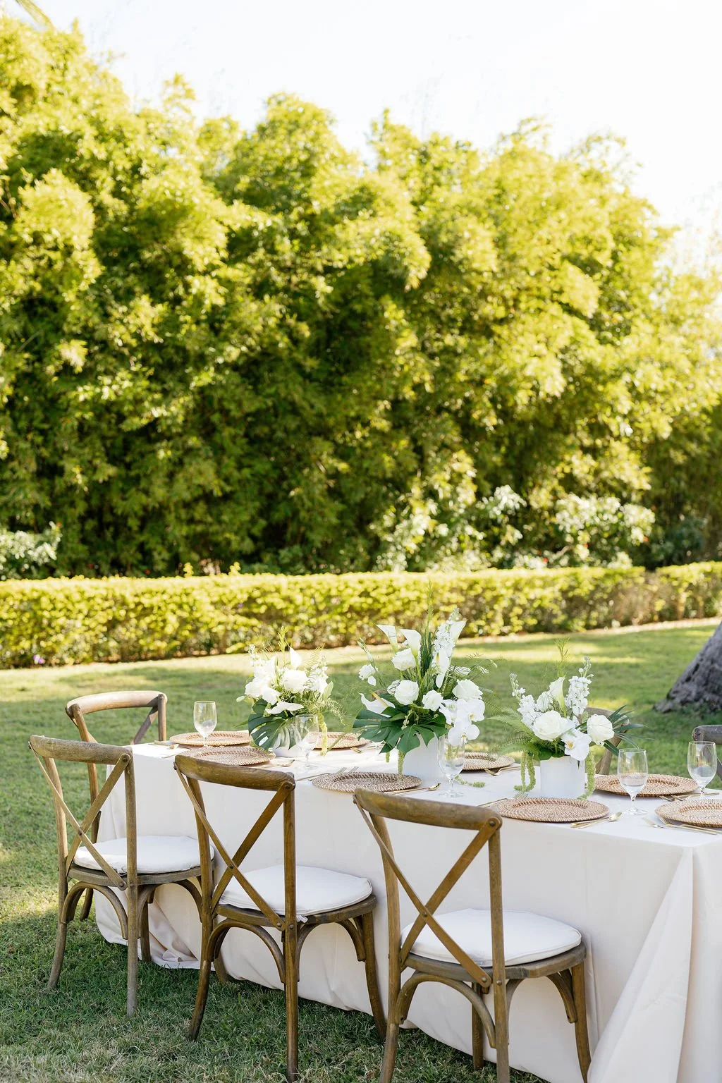Outdoor dining table set with white tablecloth, floral centerpieces, glassware, and woven placemats, surrounded by wooden chairs, in a grassy garden area with trees and shrubbery in the background.