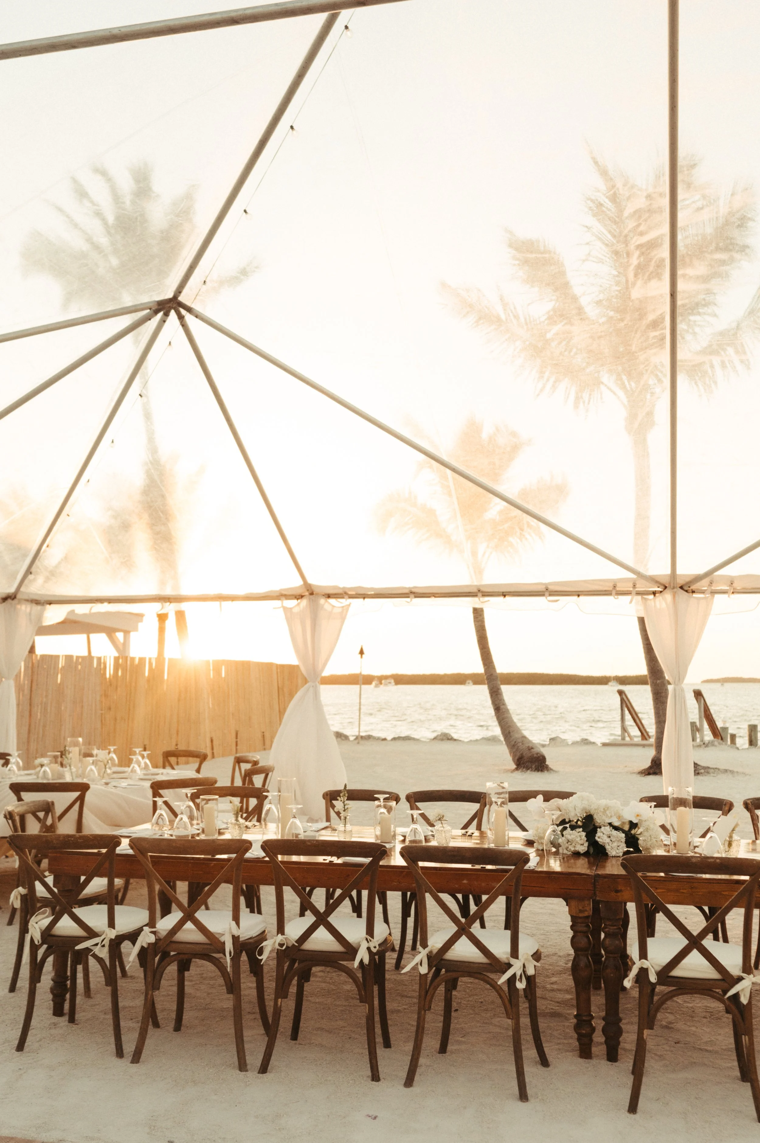 Outdoor beach setting with wooden tables and chairs under a tent, decorated for an event, with a backdrop of palm trees and a sunset over the ocean.