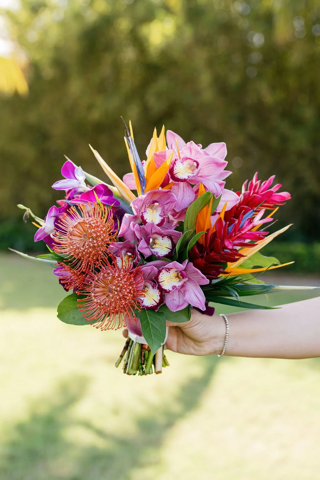 A hand holding a colorful bouquet of tropical flowers, including orchids, bird of paradise, and protea, against a blurred outdoor background.
