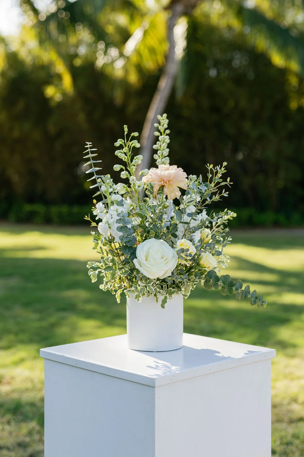 A floral arrangement with white and light pink flowers and greenery in a white vase, placed on a white pedestal outdoors with a green background and sunlight filtering through trees.