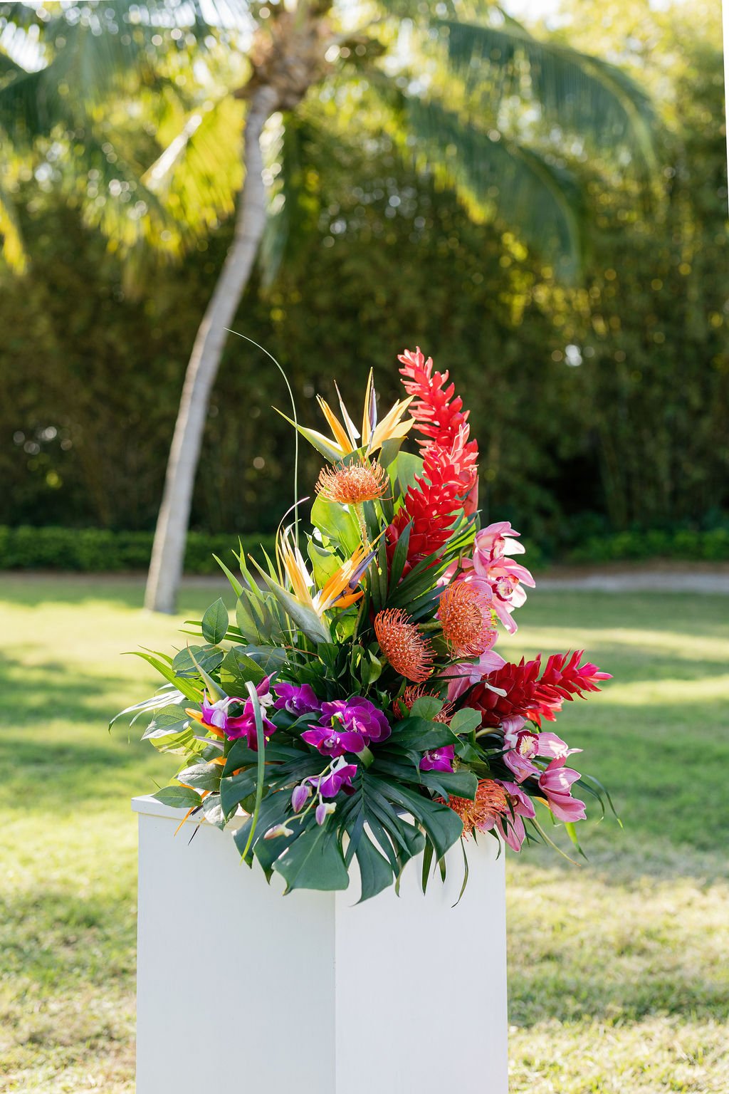 Colorful tropical flower arrangement on a white pedestal outdoors with trees and grass in the background.