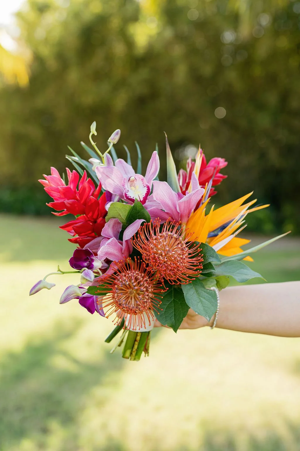 A hand holding a colorful bouquet of various flowers including pink orchids, red ginger, orange pincushion protea, and yellow and blue bird of paradise, with a blurred outdoor background.