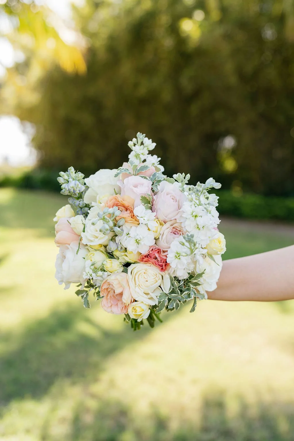 A person holding a bouquet of pastel-colored flowers outdoors, with a blurred green background.